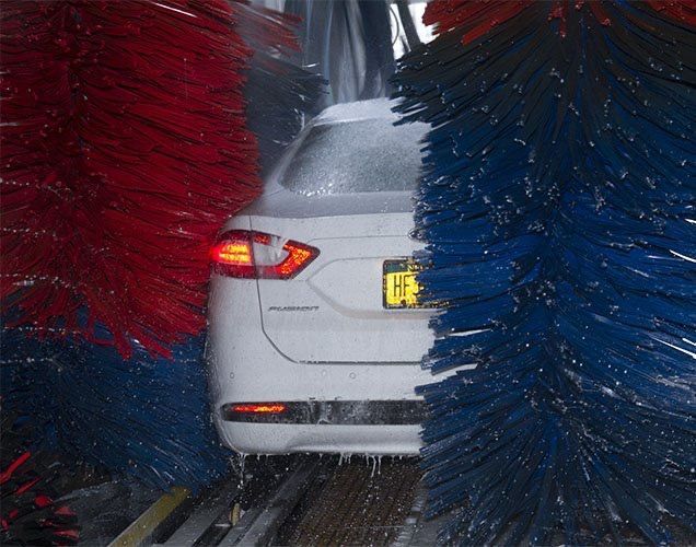 A white car passes through a car wash, framed by large, rotating red and blue cleaning brushes.