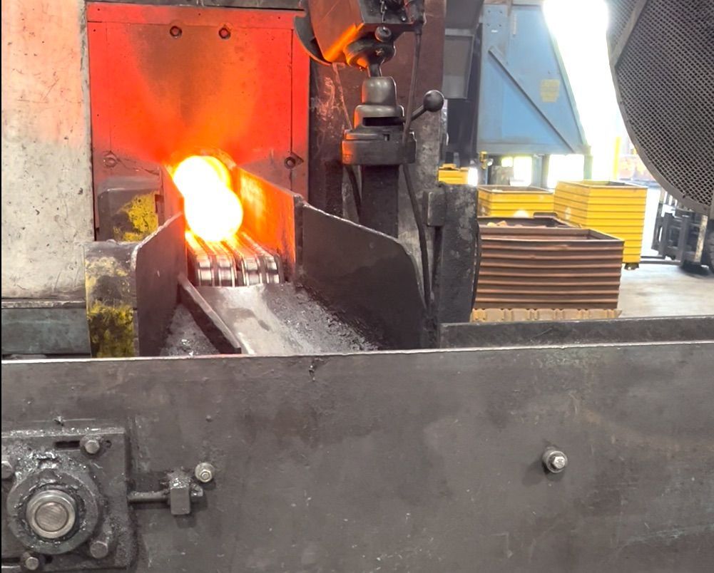 Glowing hot metal billets exit an industrial furnace onto a conveyor belt in a factory setting.