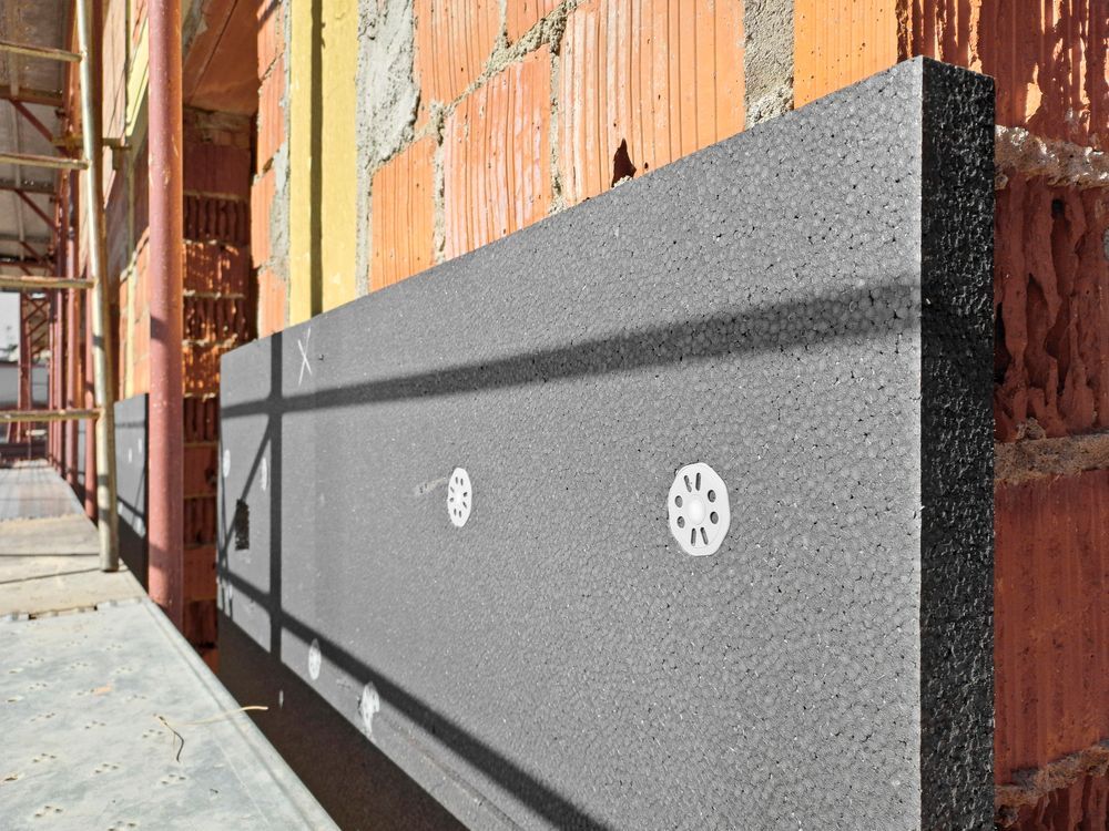 Pannello isolante grigio montato sulla facciata esterna di un edificio in mattoni non rifiniti, vicino a un'impalcatura.