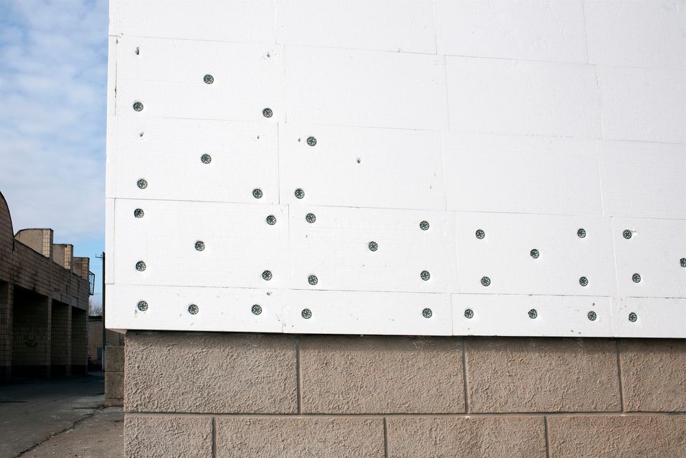 Muro bianco con file di bulloni metallici sopra una base di pietra, accanto al bordo di un edificio e al cielo azzurro.