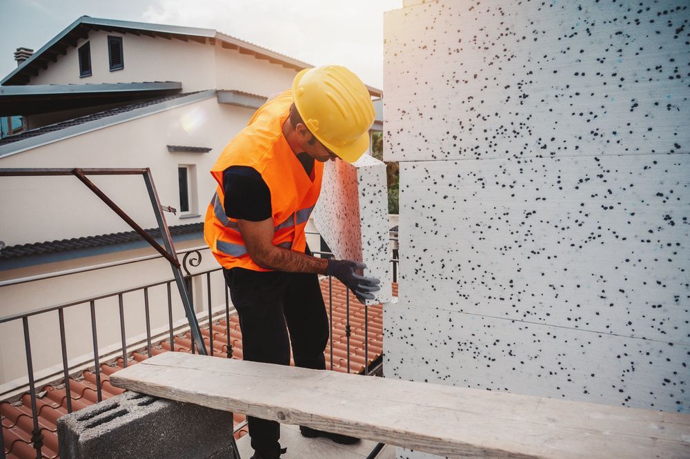 Un operaio con giubbotto di sicurezza arancione e casco giallo misura il bordo di un tetto accanto a un muro bianco.