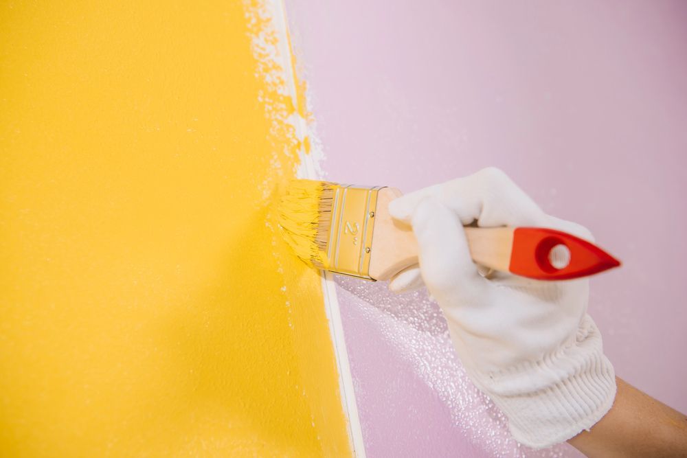 Mano guantata che dipinge con un pennello una parete di giallo accanto a una sezione rosa.