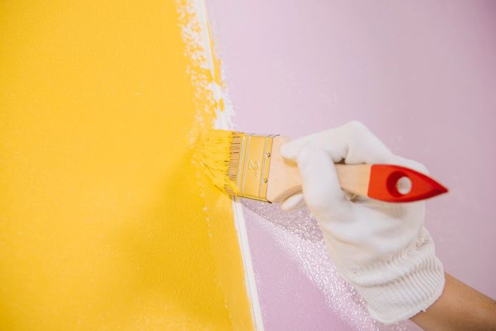 Mano guantata che dipinge con un pennello una parete di giallo accanto a una sezione rosa.