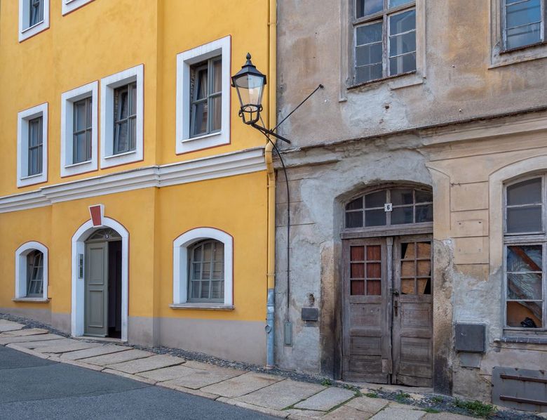 Antica strada colorata con edifici in pietra gialla e consumata dal tempo, finestre con persiane e un lampione a muro.