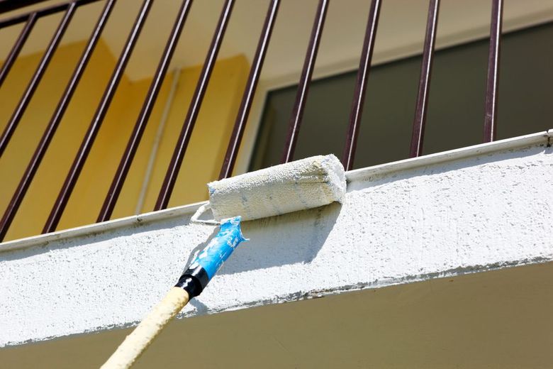 Rullo da pittura che applica vernice bianca sul davanzale e sui montanti della ringhiera di un balcone esterno.
