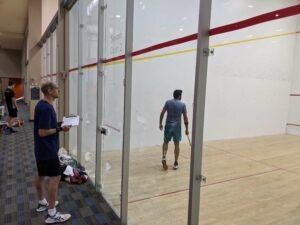 Two men are playing squash on a court behind a glass wall.