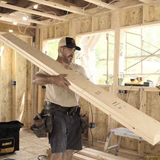 Worker carrying a long wooden beam inside a framed house under construction.