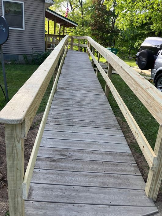 Wooden ramp with railings leading up beside a house, bordered by grass and trees.