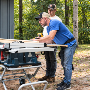 Two people using a table saw outdoors in a wooded yard, wearing safety gear.