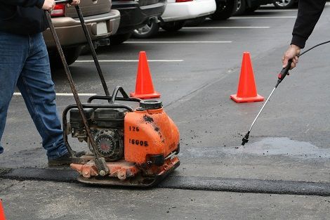 Worker using a jackhammer to repair cracked pavement beside orange traffic cones in a parking lot