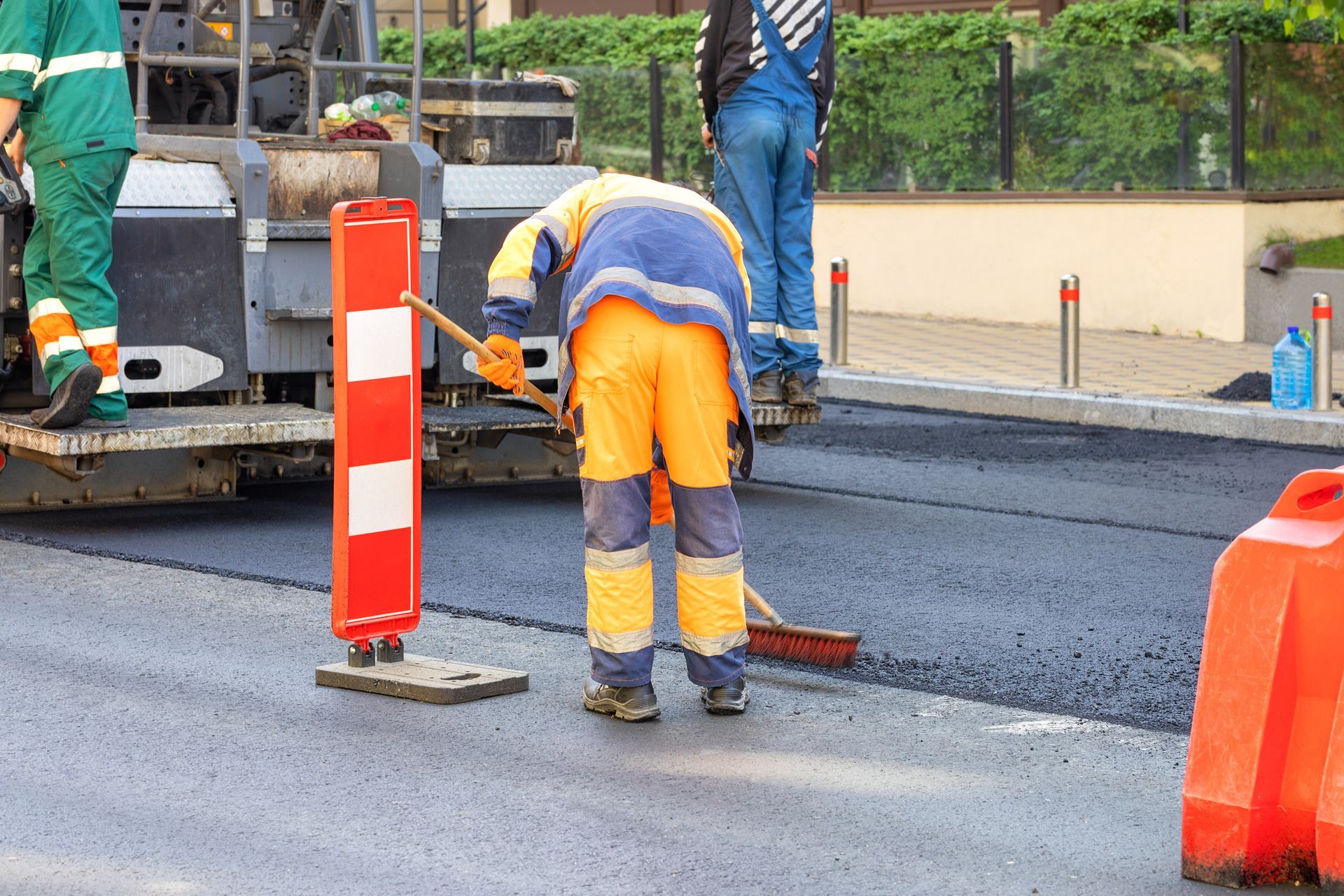 Road crew worker in reflective gear sweeping fresh asphalt paving near a paver and safety barriers.