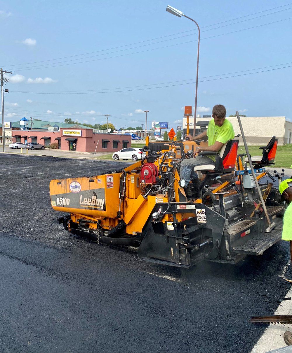 Workers operating an orange asphalt paving machine on a road construction site