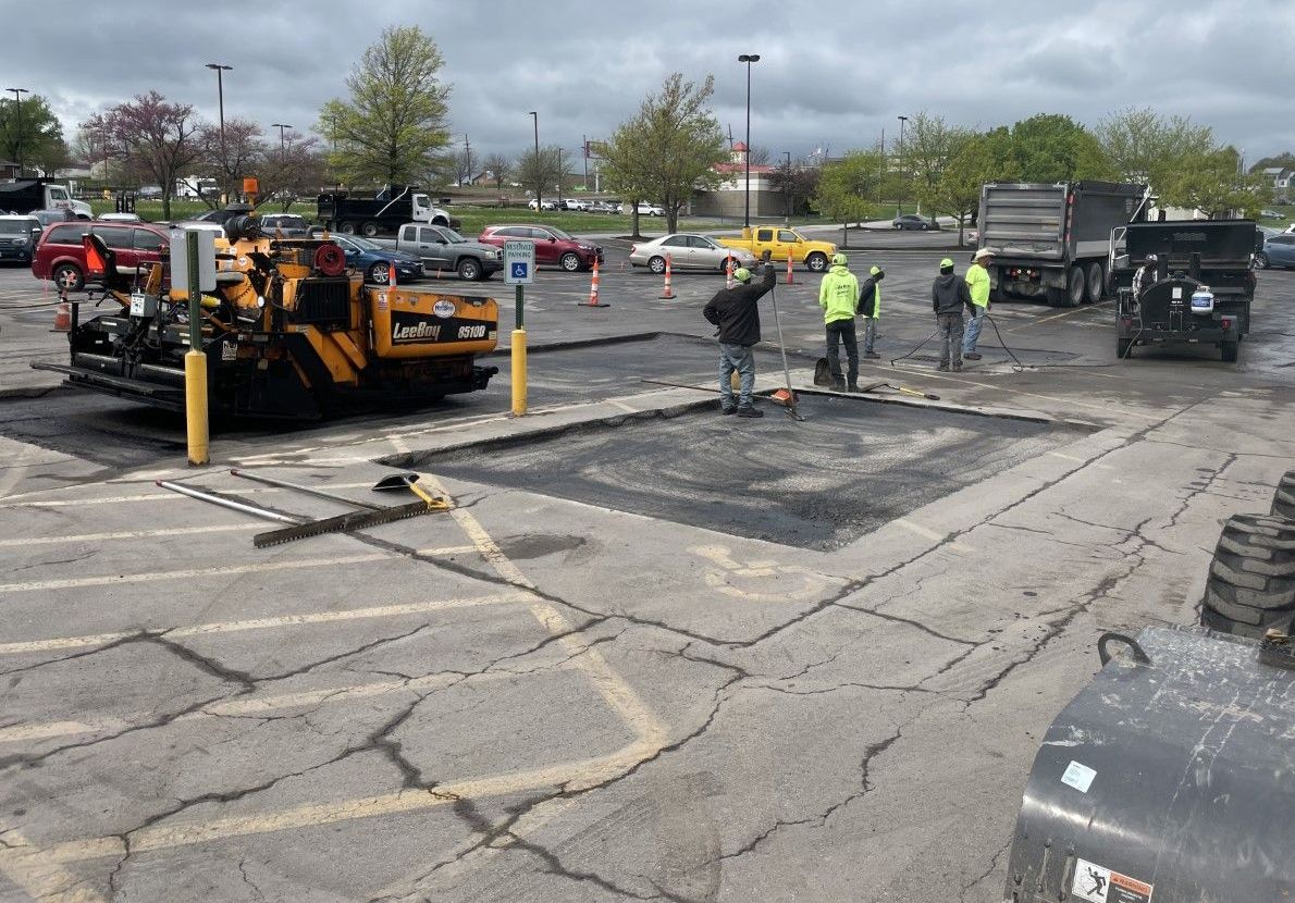Road crew paving a cracked parking lot with an orange asphalt machine and workers in safety vests