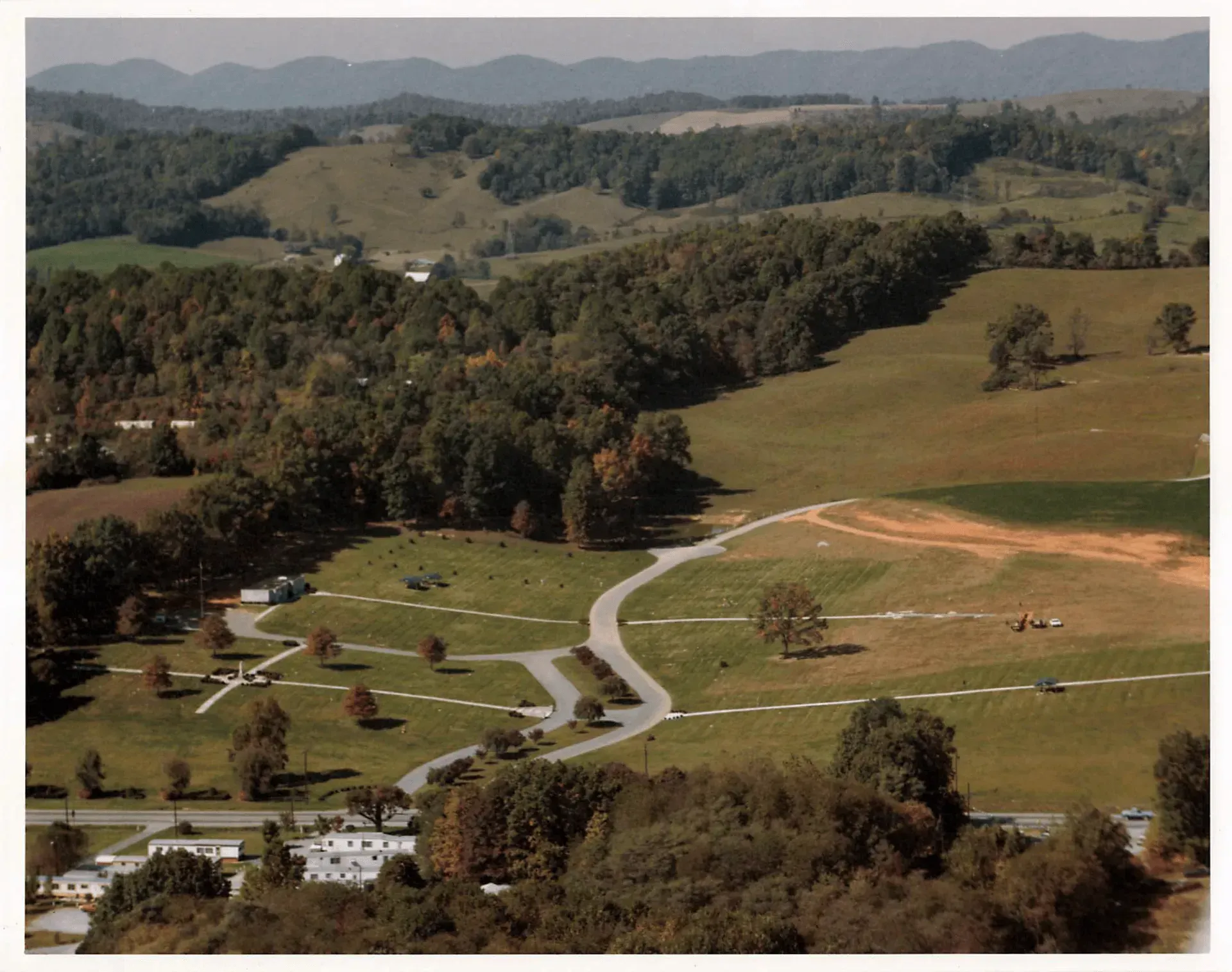 Scenic aerial view of rolling green hills, trees, and winding road, with a small building complex in the foreground.