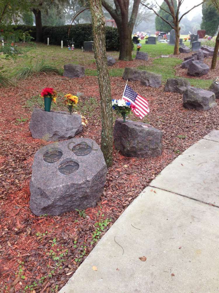 Cemetery with large granite headstones, flowers, American flag. Brown leaves and sidewalk in front of green grass.
