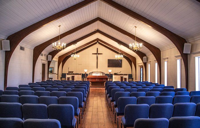 Interior of a chapel with rows of blue chairs facing a casket; a cross hangs above.