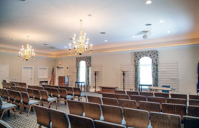 Auditorium with rows of chairs, chandeliers, windows with patterned curtains, and a podium.
