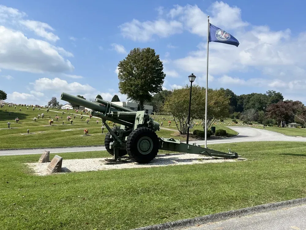 A large artillery gun on display in a cemetery, with a flag waving and headstones in the background.