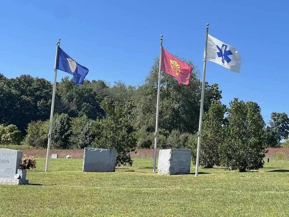 Three flags fly in a cemetery: a blue flag, a red flag, and an EMS flag, against a backdrop of trees and a blue sky.