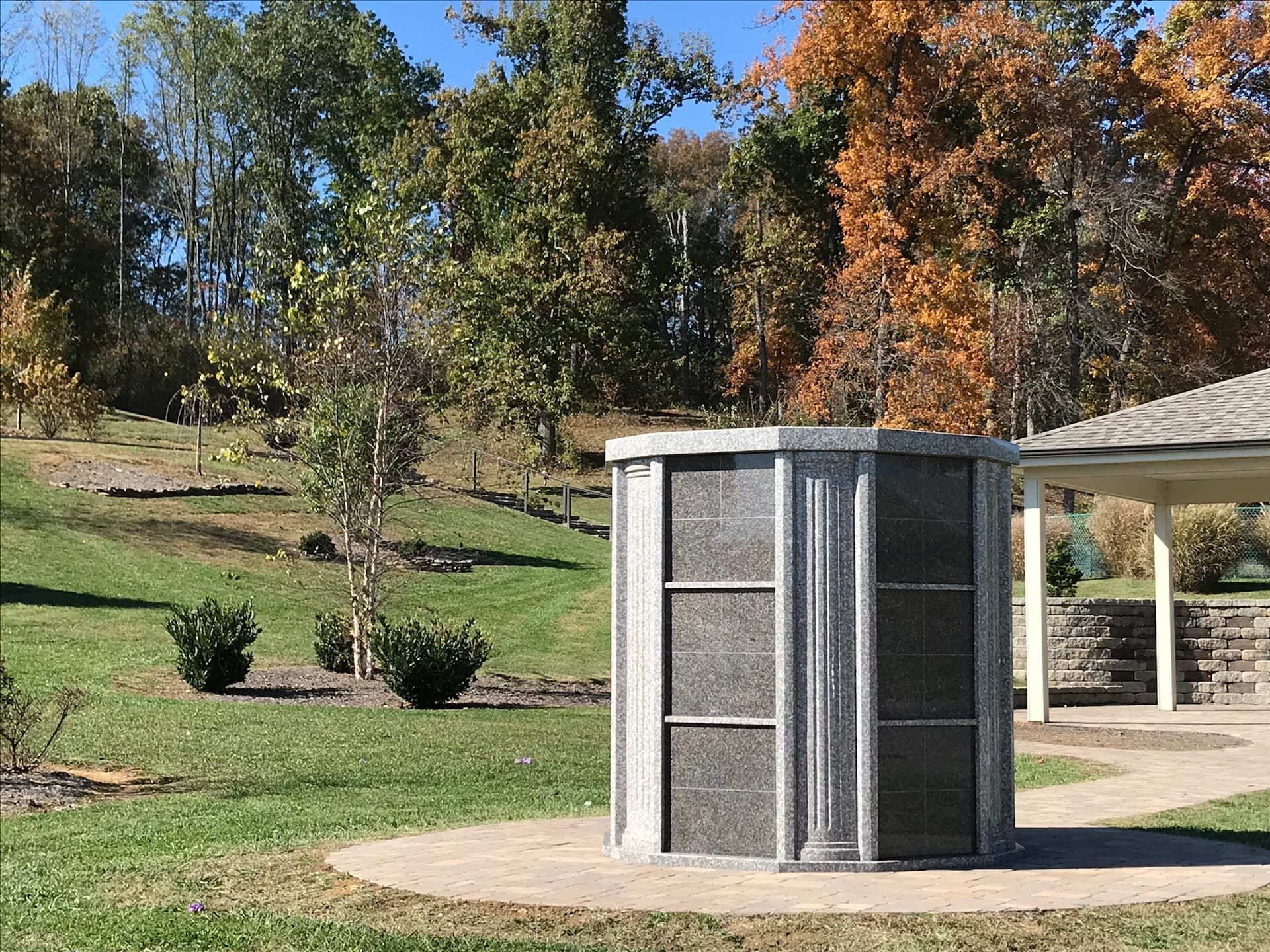 Granite columbarium with niches in a grassy cemetery setting with trees and a pavilion.