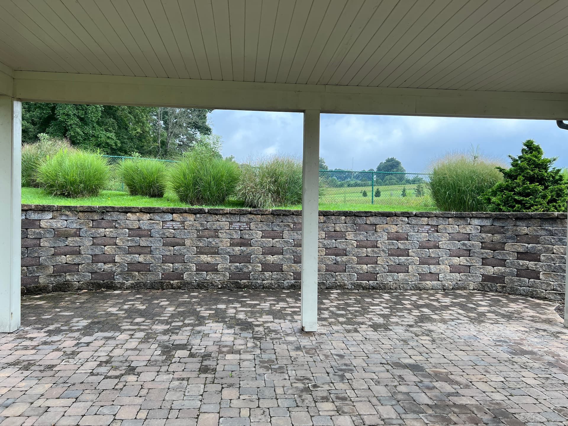 Patio with brick pavers, stone wall, green bushes, and distant trees under a cloudy sky.