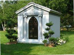 Gray mausoleum with arched door, columns, and small trimmed bushes in a grassy yard.