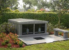 Grave with granite tomb, glass fronted niches, bench, and landscaping in a sunny cemetery.