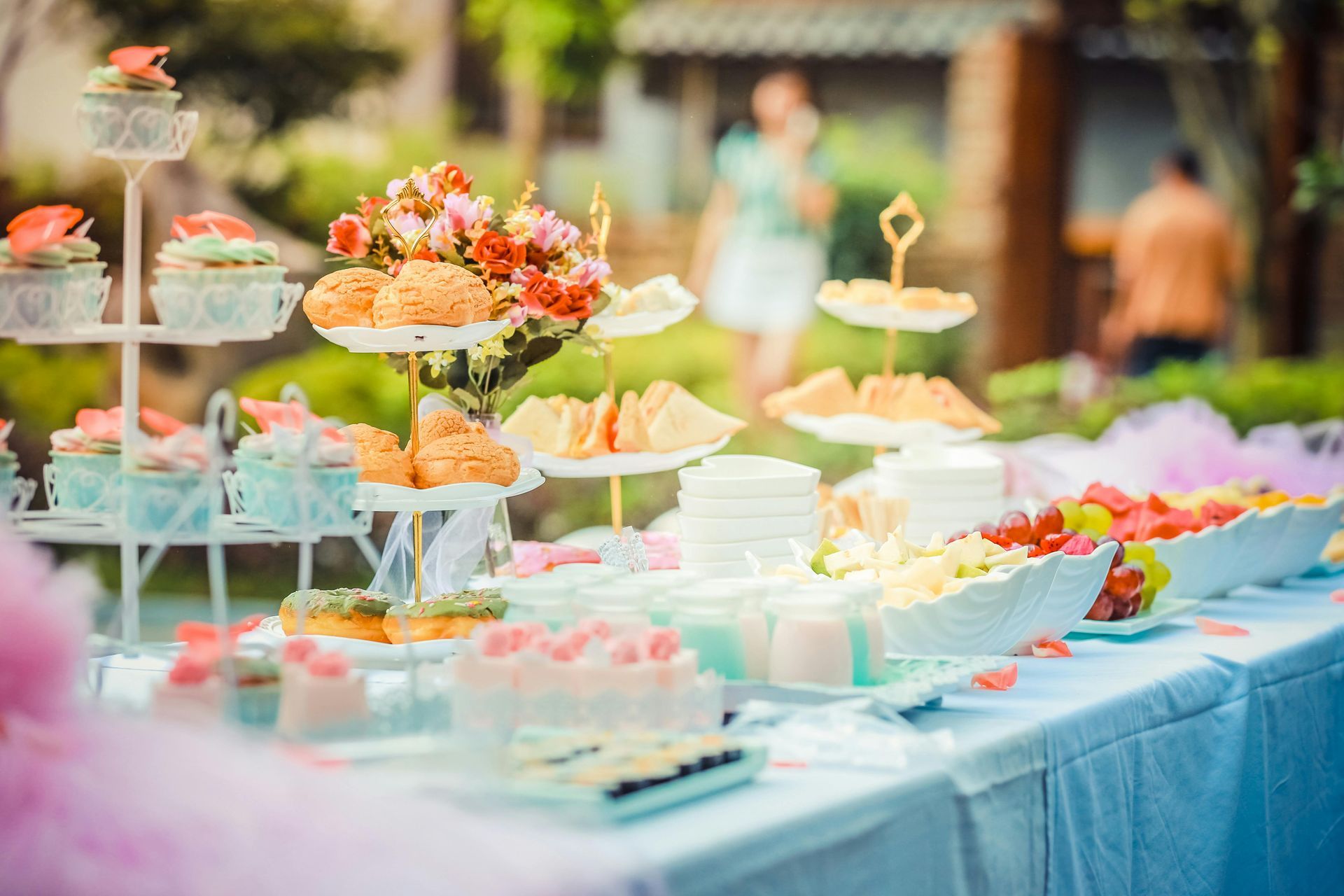 Dessert table outdoors, various pastries, cakes, and fruit. Flowers and people blurred in the background.