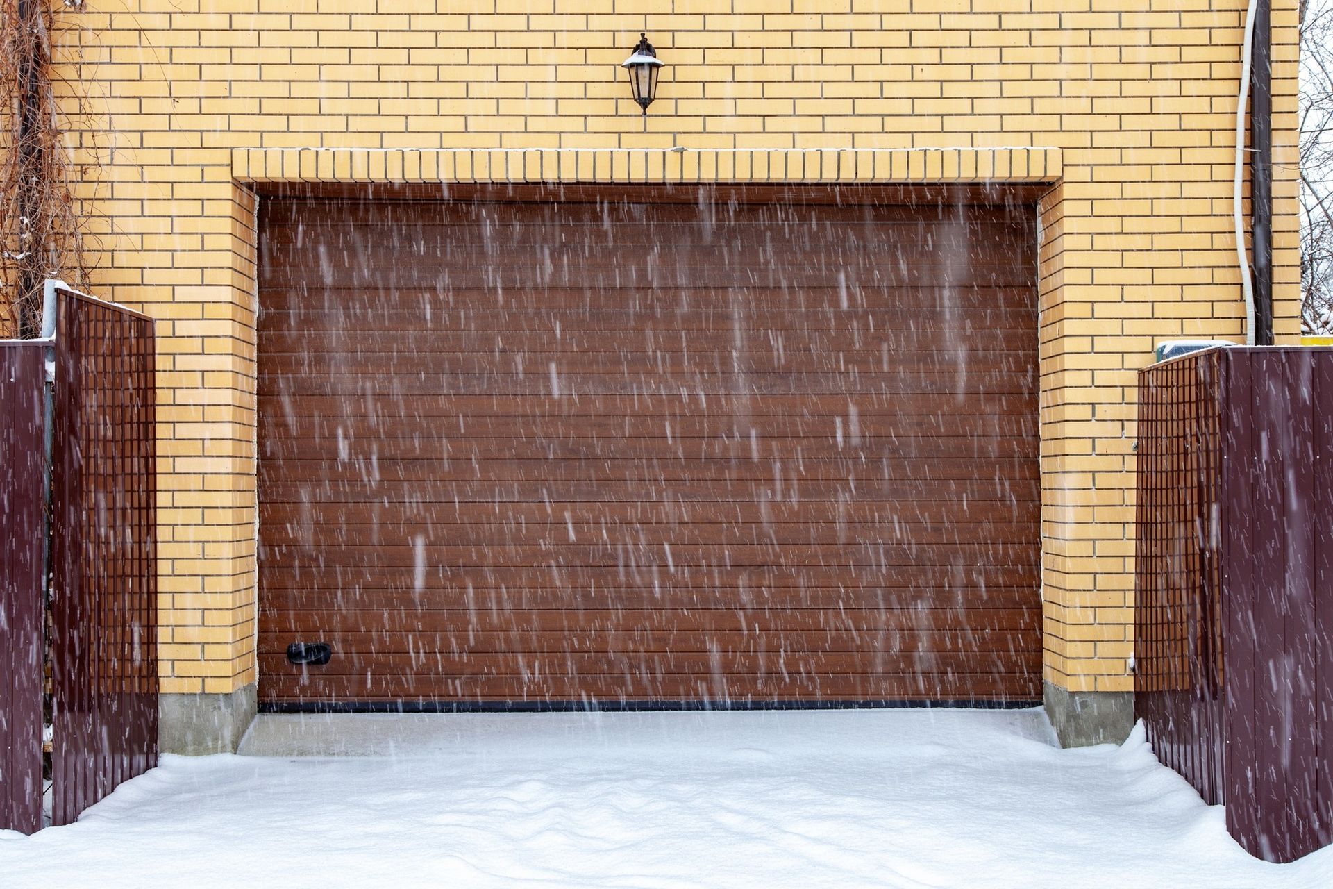 Brown garage door on a brick building, covered in falling snow. Snow on the ground.