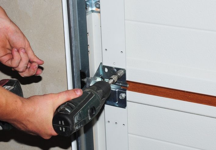 Person using a drill to install a metal bracket on a white garage door.