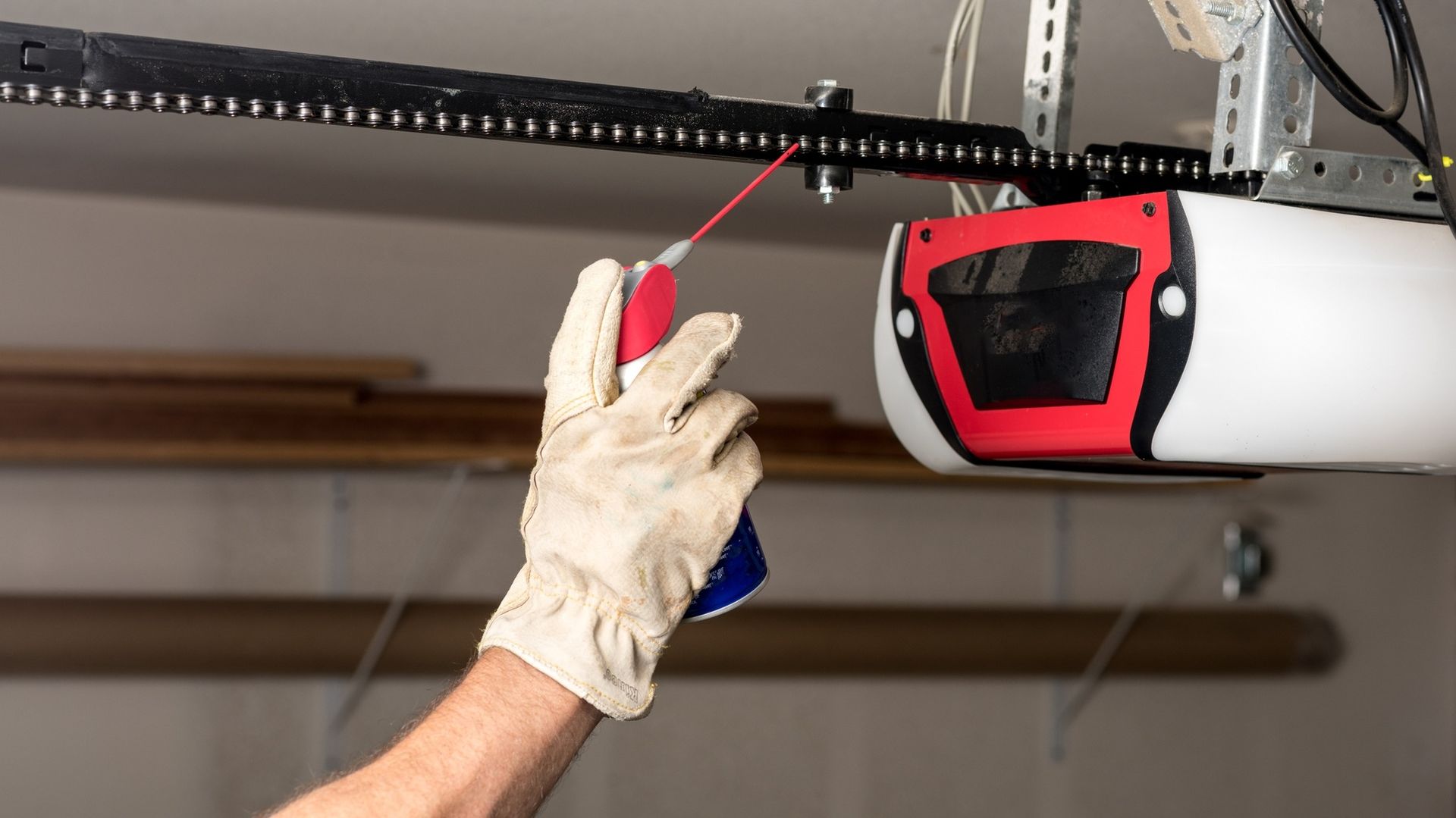 Person in gloves lubricating a garage door opener chain with a spray can.