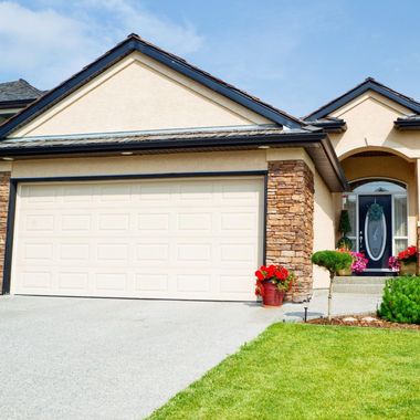 Beige house with a garage, front door, and green lawn. Red flowers accent the entrance.