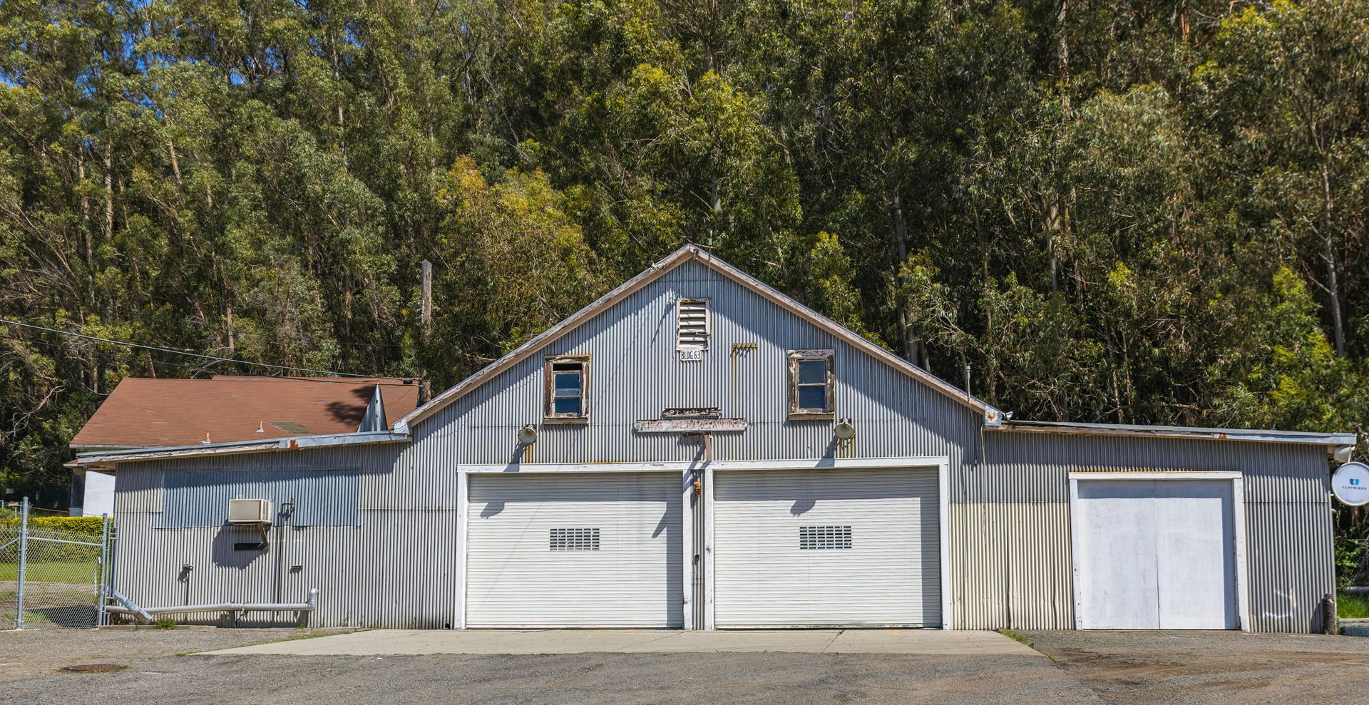 Weathered, light-gray garage with two bays, two small windows, and a small addition. Tall trees in background.