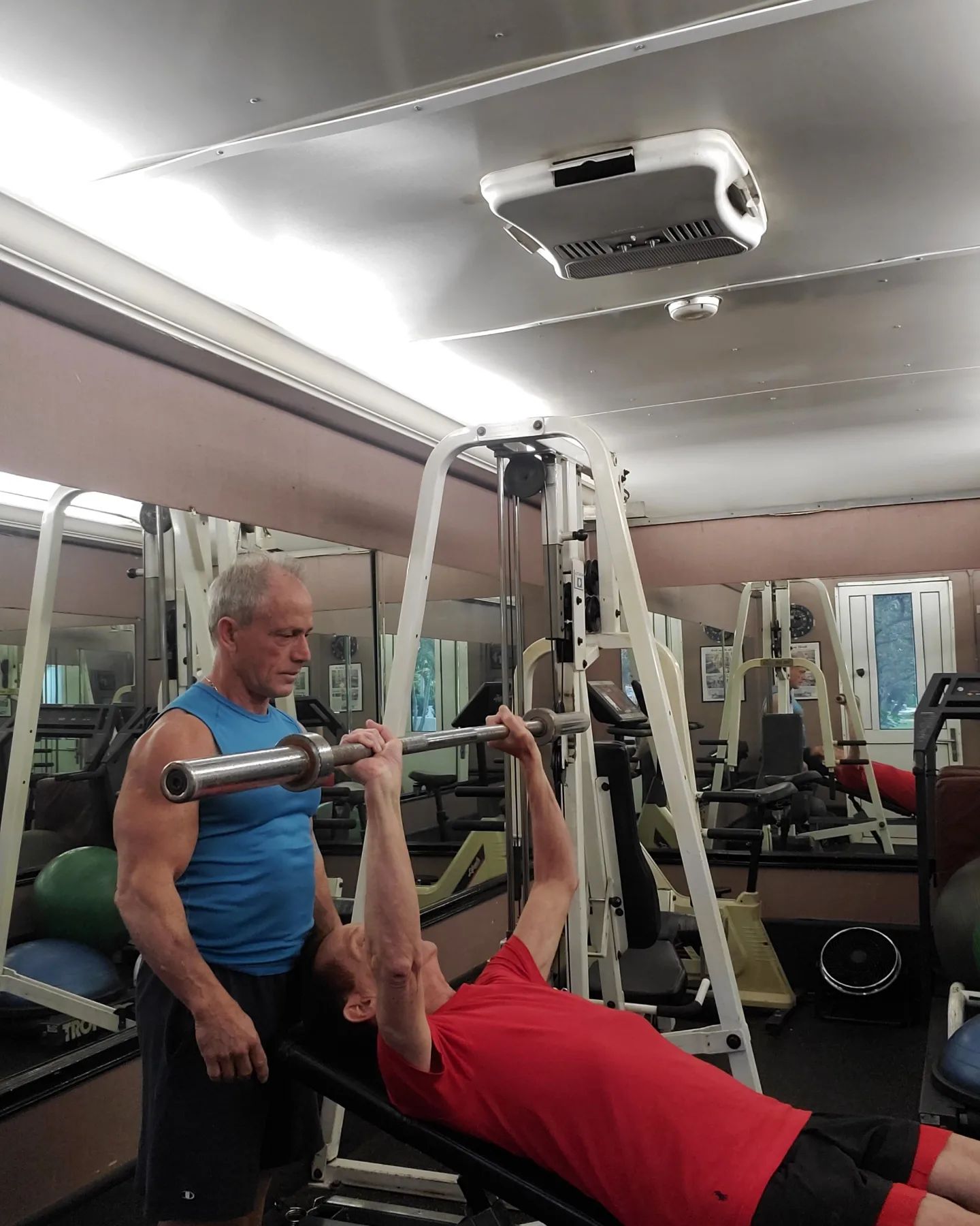 A man is lifting a barbell in a gym while another man watches.