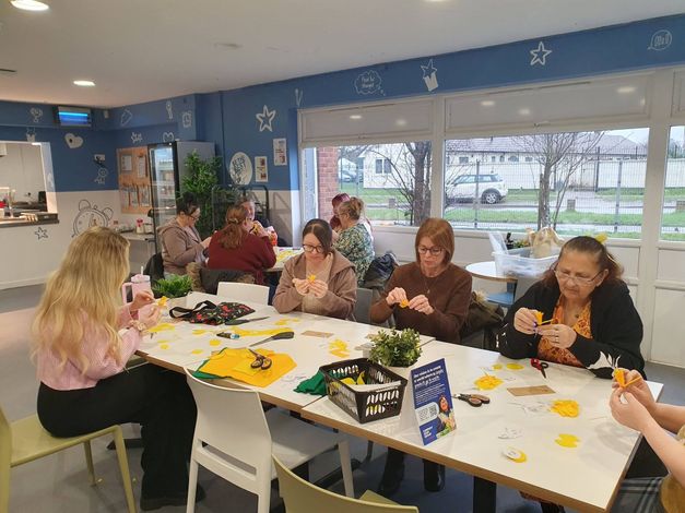 People seated around a table decorating crafts with paper, flowers, and supplies in a bright room