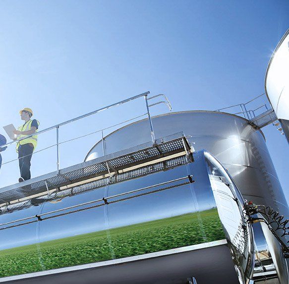 Een industriearbeider staat op een metalen tankplatform onder een helderblauwe hemel, met daaronder een reflecterende cilindrische tank.