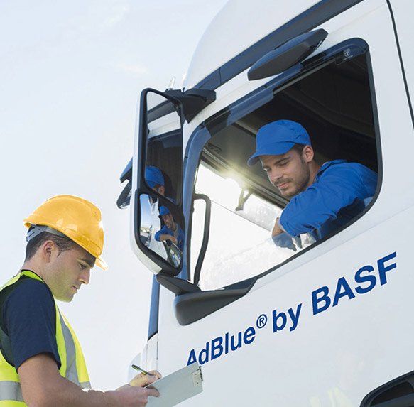 Een medewerker met een gele veiligheidshelm controleert de papieren met een vrachtwagenchauffeur met een blauwe pet bij het AdBlue by BASF-vrachtwagenraam.