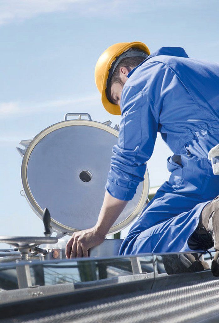 Een werknemer in een blauwe overall en een gele veiligheidshelm inspecteert industriële apparatuur in de buitenlucht.