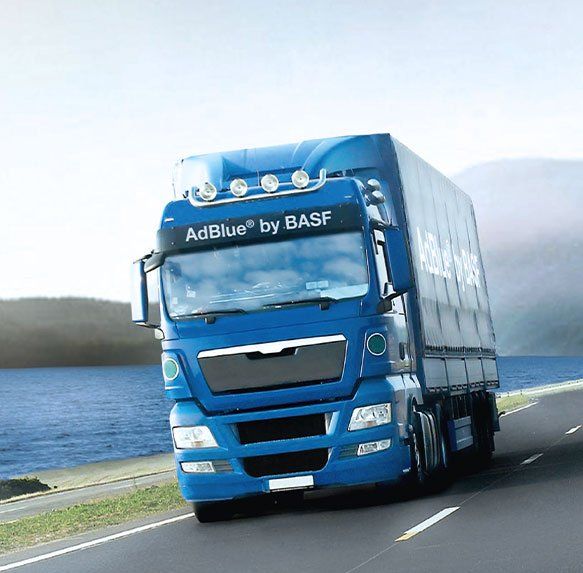 Blauwe AdBlue van een BASF-vrachtwagen die over een kustweg rijdt, langs water en heuvels.