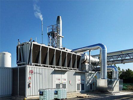 Industriële installatie met grote metalen leidingen, tanks en een schoorsteen onder een heldere blauwe hemel.