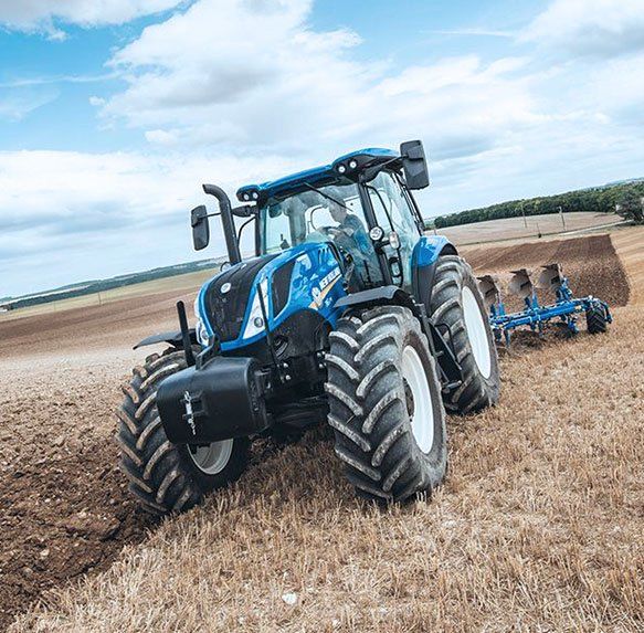 Een blauwe tractor ploegt een veld onder een bewolkte hemel, met een schijveneg erachter.