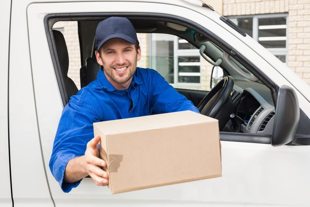 Un repartidor con uniforme azul entrega una caja de cartón desde una camioneta blanca.