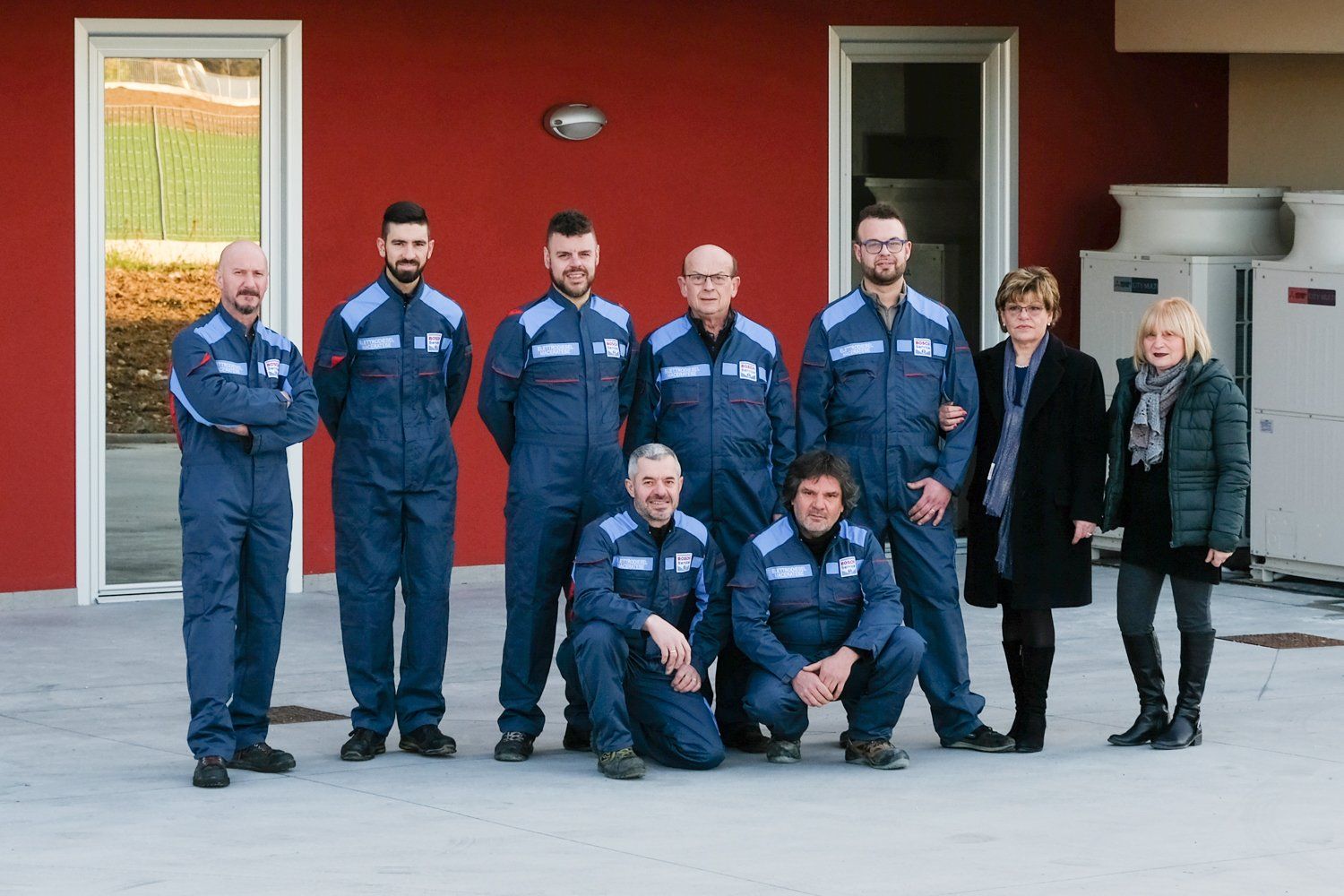 Un gruppo di persone in uniforme blu posa davanti a un edificio rosso; due donne in cappotto si uniscono al gruppo.