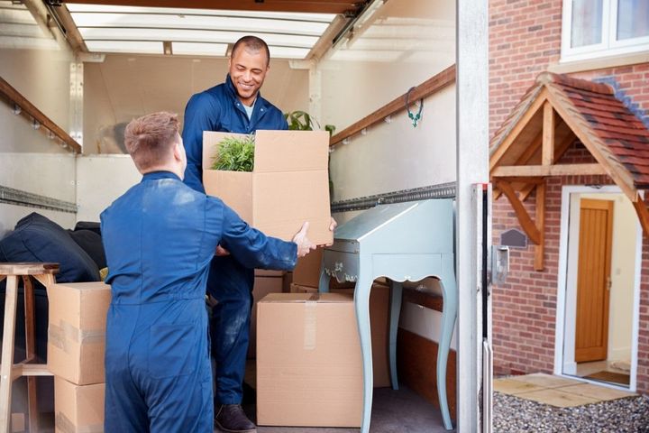 Two movers in blue coveralls carrying boxes in a moving truck, with a house entrance beside it.