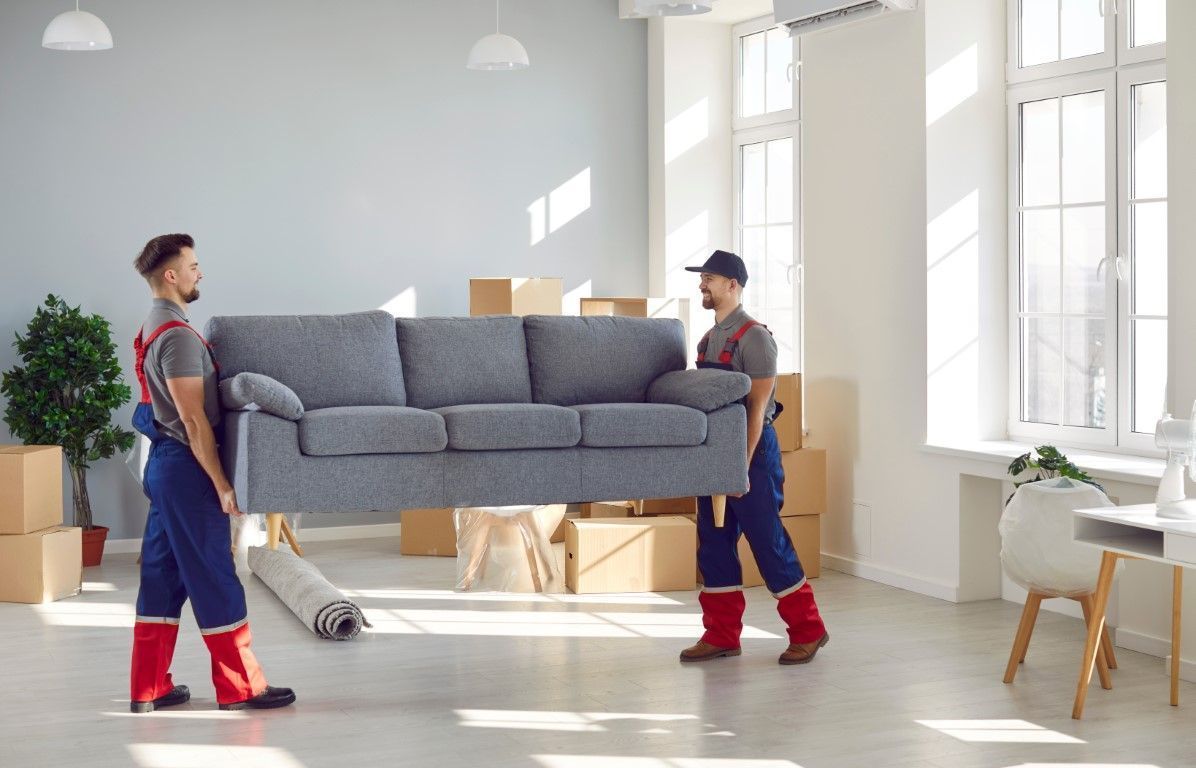 Two movers carrying a gray sofa through a bright living room with boxes and large windows