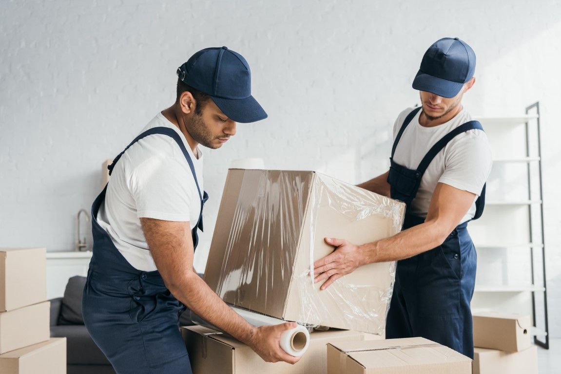 Two movers in uniforms packing a cardboard box in a bright room