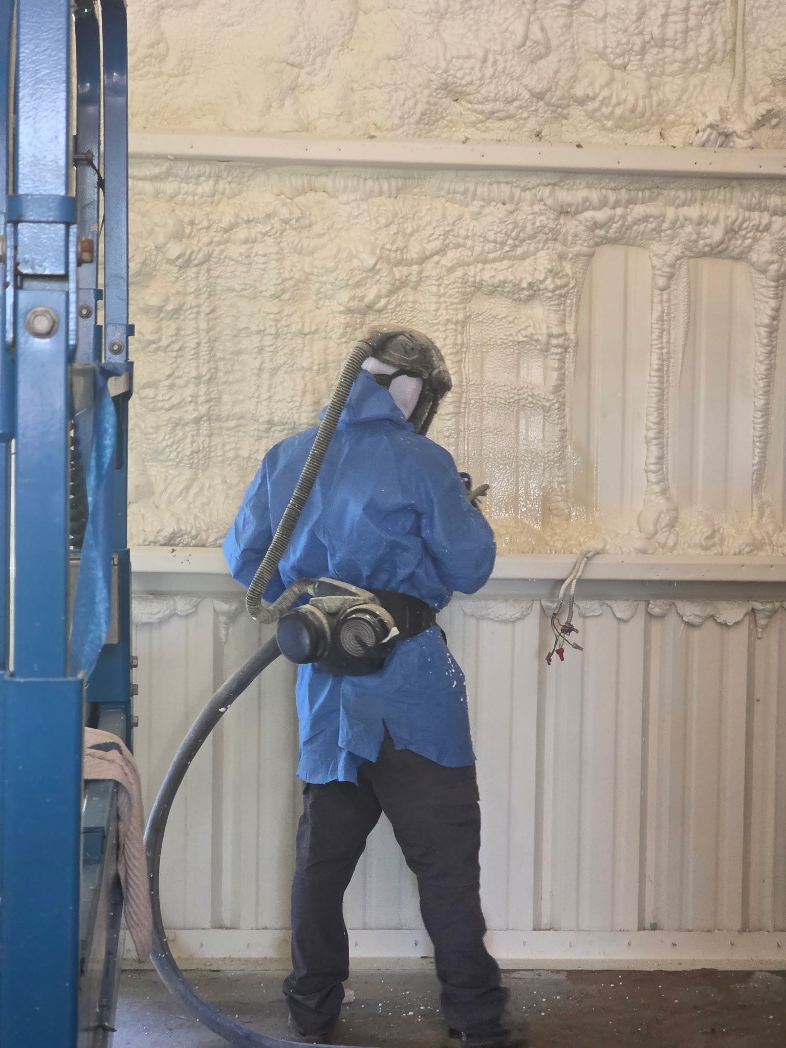 Worker in blue protective gear spray-painting a beige wall inside an industrial space