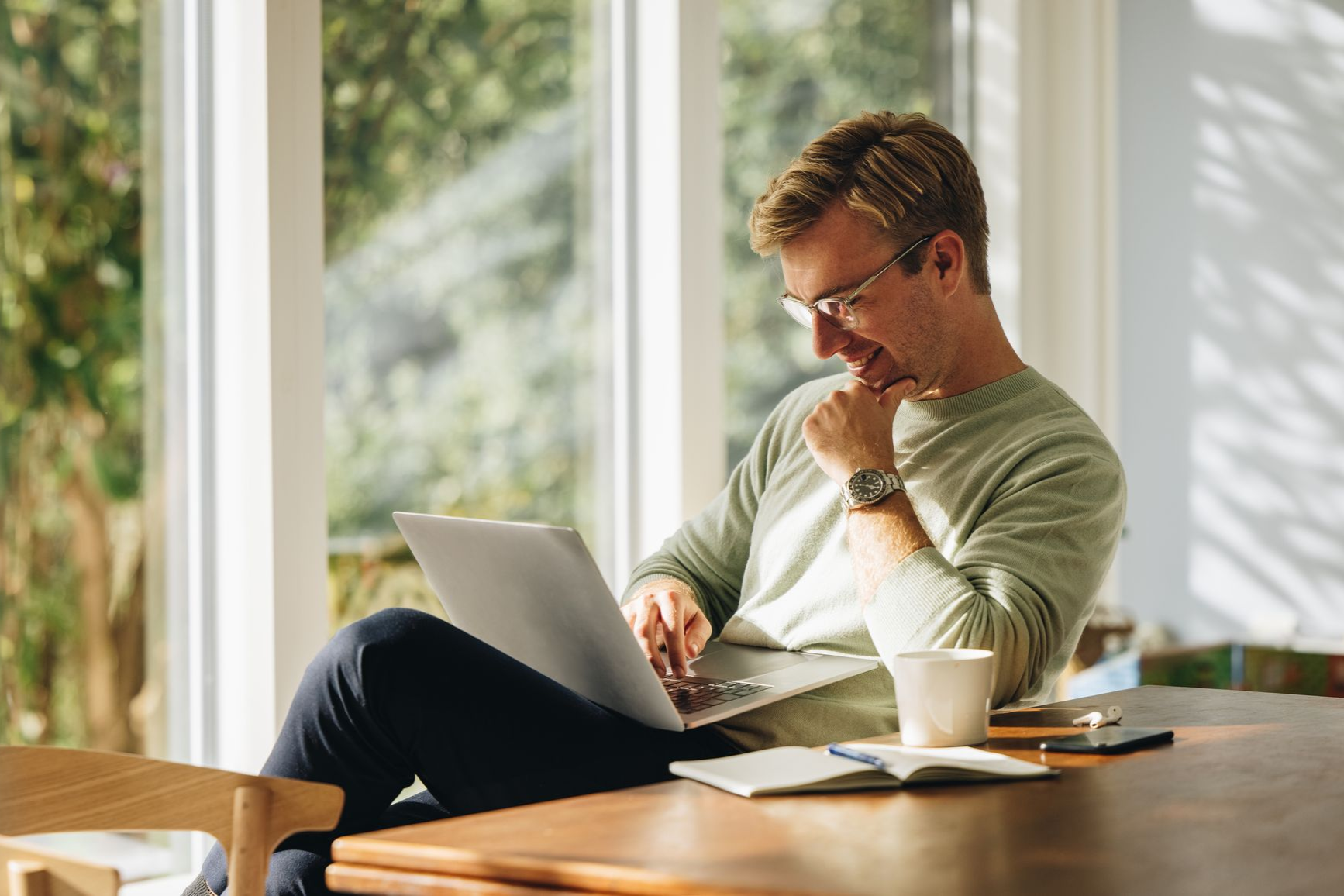 Man in glasses smiles while working on laptop at a table near a window.