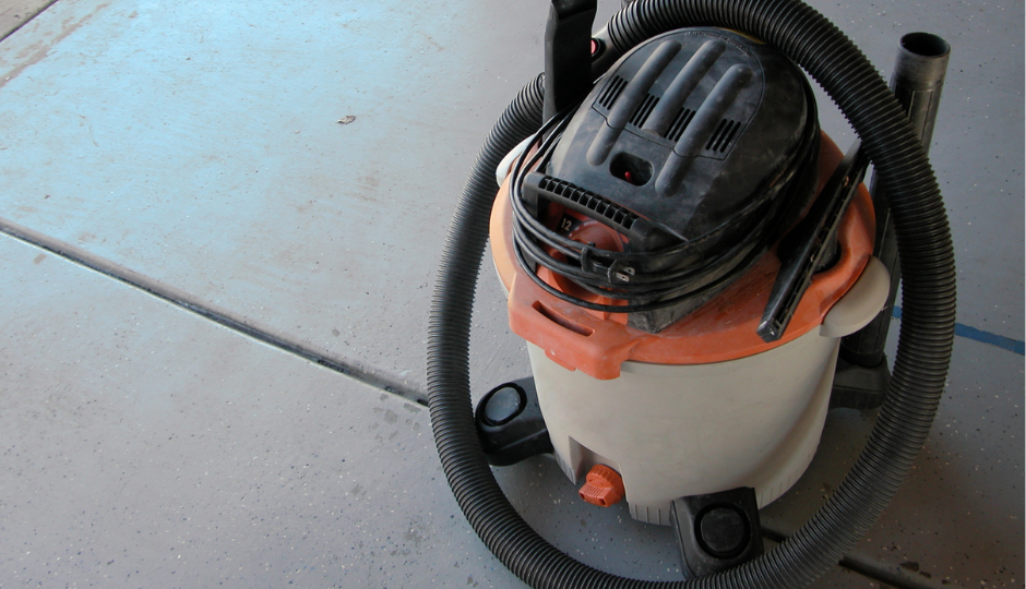 A wet-dry shop vacuum with an orange lid and a black hose, sitting on a gray concrete floor.