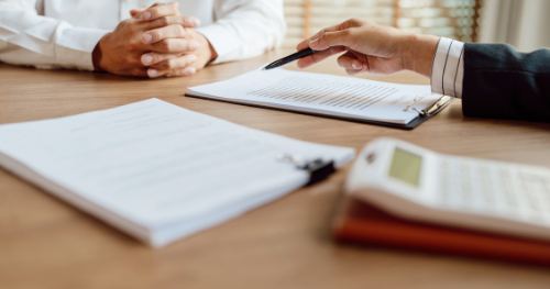 Hands clasped across a table during a business meeting with papers, notebook, and calculator visible.